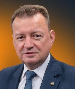 Mariusz Błaszczak przemawiający z mównicy podczas wydarzenia politycznego, gestykulujący z pewnością siebie.