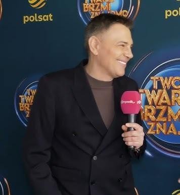 Krzysztof Ibisz w studiu telewizyjnym, energicznie prowadzący program, z mikrofonem w dłoni i gestykulujący, na tle ekranu z grafiką teleturnieju.