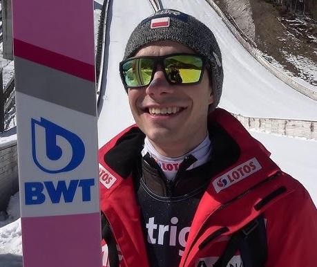Jakub Wolny w locie podczas skoku narciarskiego, z widocznym ośnieżonym stokiem w tle.